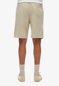Pantaloni corti in cotone beige con cintura elastica, tasca posteriore e cuciture in vista. Indossati con sneaker beige chiaro e calze grigie.