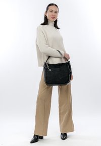 Sieviete, tērpusies bēša krāsas augstu uzpildītā džemperī un biksēs, turot melnu rokassomiņu, stāvot pret vienkāršu balto fonu.