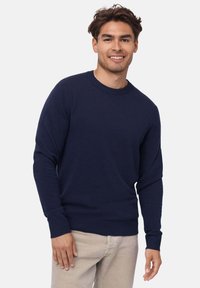 Pullover blu navy realizzato in tessuto morbido, con maniche lunghe e polsini a coste. Indossato con pantaloni beige chiaro.