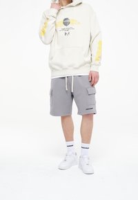 Beige mikina s žlutým grafickým designem a žebrovaným lemem, kombinovaná se šedými cargo šortkami s bočními kapsami a bílými teniskami.