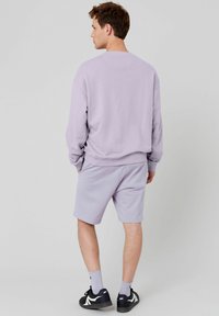 Ensemble en sweatshirt et shorts lilas, fabriqué en tissu doux. Le sweatshirt a une coupe décontractée, des poignets côtelés, et les shorts sont dotés de poches. Baskets noires.