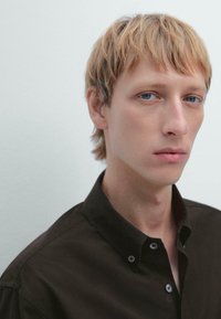 Giovane con capelli biondi corti e pelle chiara, indossa una camicia scura con bottoni, guarda leggermente di lato su uno sfondo neutro.