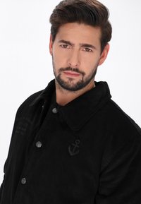 Fekete corduroy dzseki nagy gallérral, fémből készült gombokkal és horgony emblémával. A textúrált anyag látható bordákkal és varrási részletekkel rendelkezik.