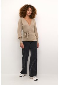 Suéter envolvente beige con un profundo escote en V, mangas largas abullonadas y cinturilla con lazo. Combinado con pantalones de pierna ancha en gris oscuro de tiro alto y zapatillas blancas.