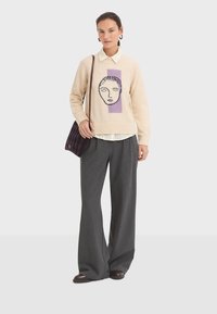 Knit sweater in beige with a graphic face design in purple and black. Paired with wide gray pinstriped trousers and a striped bag.