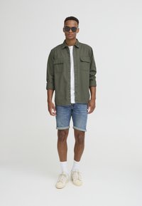 Olive green button-up shirt over a white t-shirt, paired with denim shorts and cream sneakers. Model wears dark sunglasses and white socks.