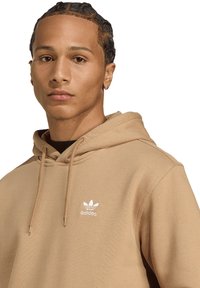 Tan-kleurige hoodie van zachte stof, met een capuchon met treksluiting en een wit Adidas-logo op de borst. Strakke lijnen en een aansluitend ontwerp.