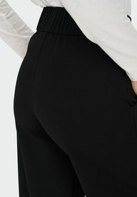 Sorte tapered bukser med elastisk talje, glat tekstur og synlige sidelommer, vist fra en let sidevinkel.