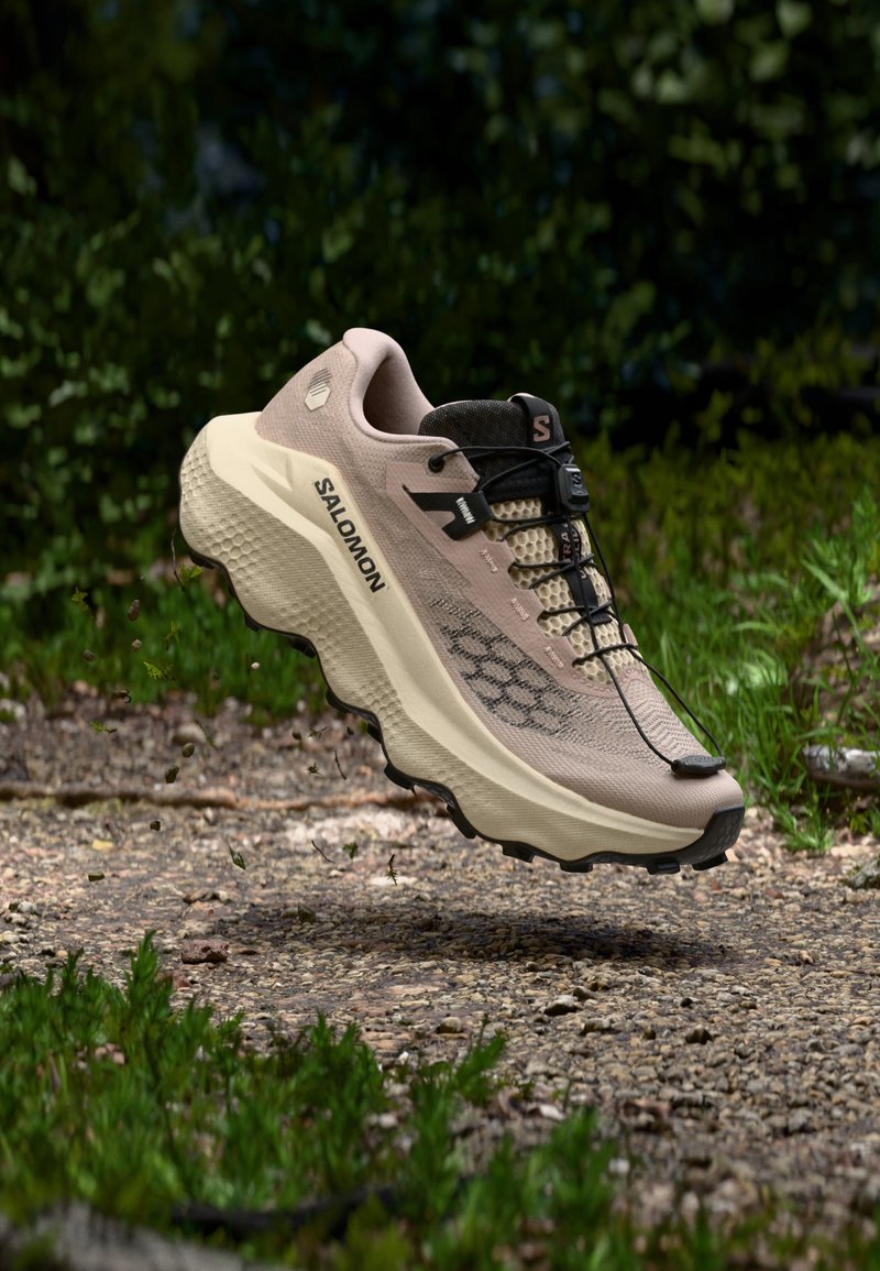 Chaussure de trail Salomon beige et noire avec semelle robuste flottant au-dessus d'un chemin de terre avec un fond de feuillage vert.