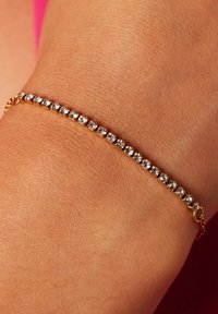 Bracelet en ton or avec des strass ronds transparents disposés en motif linéaire, porté au poignet sur un fond de peau.