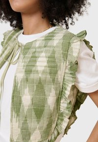 Gilet verde e crema con un motivo checkered testurizzato e bordi arricciati. Si lega al colletto, indossato sopra una t-shirt bianca.