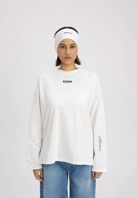 Biała koszula z długim rękawem z czarnym logo na klatce piersiowej i tekstem na rękawie, w połączeniu z jasnoniebieskimi luźnymi dżinsami. Biała opaska na głowę.