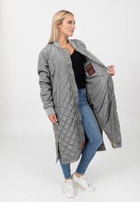 Grijze gewatteerde lange jas met geribde manchetten en een gouden ritssluiting. Binnenlabel is zichtbaar. Model draagt een zwarte top en blauwe jeans met witte sneakers.