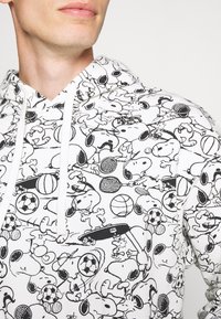 Felpa con cappuccio bianca con stampa nera di Snoopy in stile cartone animato e vari attrezzi sportivi. Il materiale sembra morbido; il design è continuo e dettagliato.