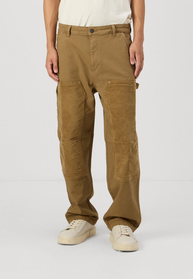 Pantalons cargo marron avec des patches en velours côtelé texturé, cinq poches et une coupe décontractée, associés à des baskets claires.