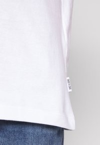 Bílý bavlněný tričko s lemem a malou bílou textilní štítkou s nápisem "ONLY & SONS" nad modrými džíny.