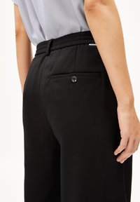 Schwarze Hosen mit glatter Textur, ausgestattet mit einer Gesäßtasche mit Knopfverschluss und einem elastischen Bund für zusätzlichen Komfort.