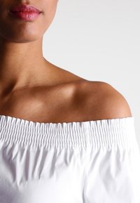 Top bianco off-the-shoulder con smock texture sul colletto e tessuto liscio, che rivela una spalla e parte del collo.