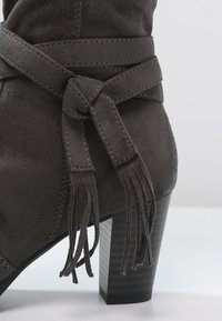 Bottine en daim gris foncé avec un talon bloc et une lanière décorative nouée ornée de franges autour de la partie supérieure de la cheville.