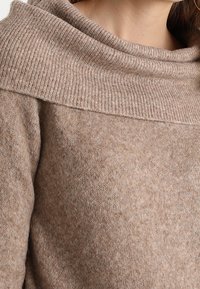 Pull en laine beige avec un col bénitier large et côtelé ainsi qu'un corps doux et texturé. Présente une teinte subtile et chaleureuse ainsi qu'une coupe décontractée.