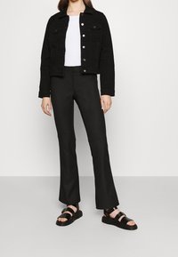 Mustat farkkutakki lyhyellä leikkauksella ja hopeisilla napeilla, yhdistettynä pitkään mustaan flare-housuun ja mataliin mustiin sandaaleihin.