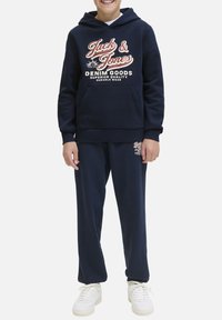 Sweatshirt marine avec capuche et poche frontale, arborant un texte graphique blanc et rouge, associé à un jogging marine assorti et des baskets blanches.