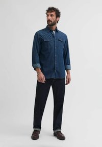 Tumši zilo denimu krekls ar divām krūšu kabatām, snap pogām un sarullētām piedurknēm, apvienots ar tumšiem džinsiem un brūnām kurpēm. Vienkārša balta fona.