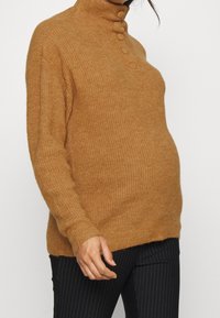 Femme portant un pull en maille couleur chameau avec des boutons recouverts de tissu sur un col montant et un pantalon noir à fines rayures.