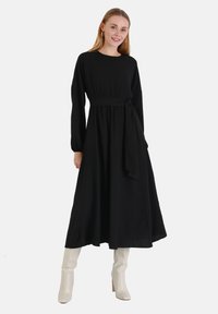 Schwarzes langärmeliges Kleid mit tailliertem Schnitt und fließendem Rock, ausgestattet mit einem gebundenen Gürtel. Glattes Material, minimalistisches Design und knöchelhohe weiße Stiefel.