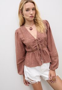 Brun linblus med broderade detaljer, knytkrage och puffärmar, som parats med vita slitna shorts och ett cirkulärt halsband.