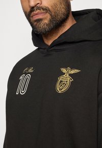 Crna dukserica s zlatnim grbom orla i natpisom "O Rei 10" sprijeda. Ima mekanu teksturu i klasični dizajn s kapuljačom.