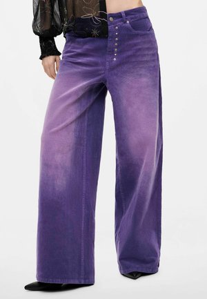 Persoon draagt een paarse fluwelen broek met hoge taille, wijde pijpen en knopen, gecombineerd met een doorschijnende zwarte blouse met bloemenmotief, staand op een witte achtergrond.