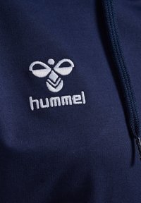 Tamsiai mėlynas džemperis su lygiu paviršiumi, turintis baltą siuvinėtą logotipą ir tekstą "hummel" krūtinės srityje. Apima suveržimo gobtuvą.