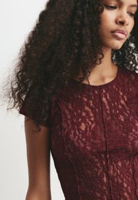 Junge Frau mit langen, lockigen Haaren, die ein kurze Ärmels burgunderfarbenes Spitzenoberteil trägt und mit neutralem Ausdruck auf einen einfarbigen Hintergrund schaut.