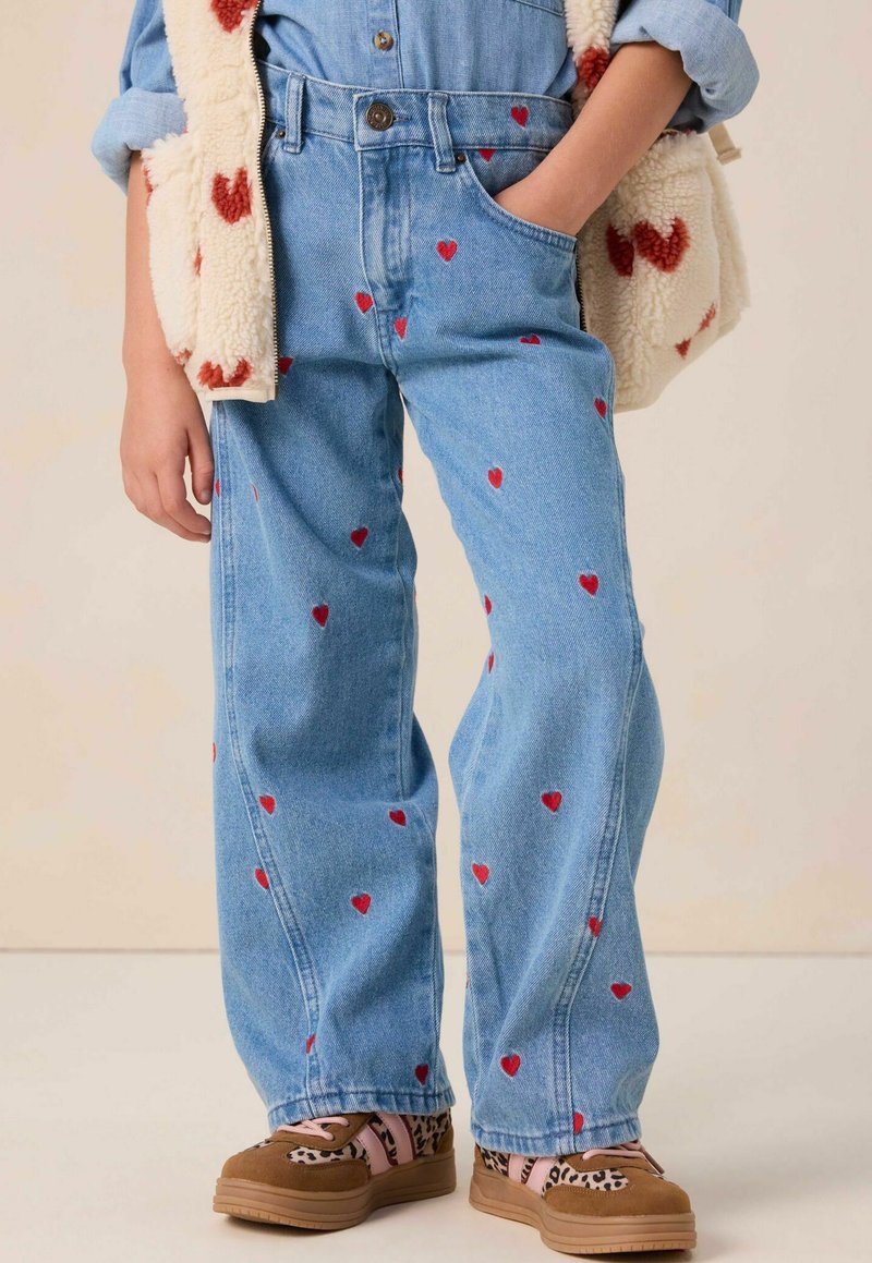 Jean en denim bleu avec broderie en forme de cœur rouge, coupe décontractée et taille haute. Associé à des sneakers à motifs marron et rose.