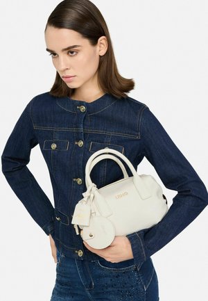 Borsa a mano bianca in pelle con due manici arrotondati, dettagli dorati, logo e un charm removibile, tenuta da una persona in una giacca di jeans blu.