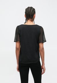 Μαύρη μπλούζα με διαφανείς, φουσκωτές μανίκια, με στρογγυλή λαιμόκοψη και λεία υφή υφάσματος. Ο σχεδιασμός στην πλάτη προσφέρει μια καθαρή, απλή όψη.