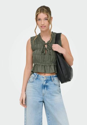 Mladá žena v olivovo zelenej topánke bez rukávov a svetlo modrých džínsoch, nesúca čiernu tašku cez rameno, stojaca pred jednoduchým pozadím.