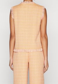 Tweed bezrukávový top v pastelově oranžové barvě s texturovaným třepení na výstřihu a lemu. Pohled zezadu, spárováno s odpovídajícími tweedovými kalhotami.
