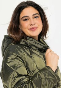 Olivgrön pufferjacka med ett kviltat mönster, hög krage och dragkedja. Materialet verkar glänsande med en slät textur.