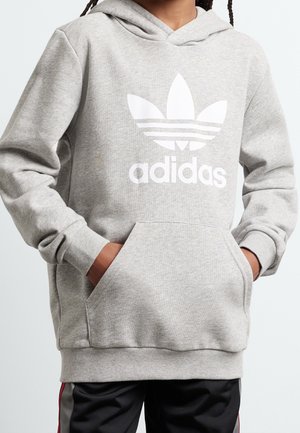 Kind trägt eine hellgraue Adidas-Kapuzenjacke mit einer Fronttasche und Logo, Hände in den Taschen, kombiniert mit schwarzen und roten Trainingshosen.