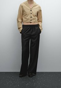 Person wearing a beige cropped buttoned jacket and loose black pants standing with hands in pockets on speckled dark floor.