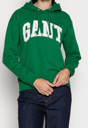 Personne portant un sweat à capuche vert avec un texte blanc "GANT", un jean bleu foncé, les mains jointes près de la taille sur un fond uni.
