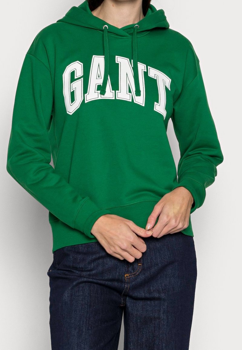 Personne portant un sweat à capuche vert avec un texte blanc "GANT", un jean bleu foncé, les mains jointes près de la taille sur un fond uni.