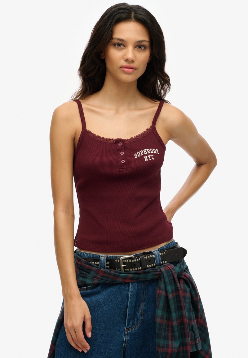 Burgundy ribbed tank top with lace trim and button detailing, featuring a "Superdry NYC" logo in white. Worn with high-waisted jeans and a plaid shirt.
