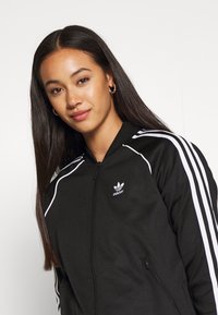 Chaqueta negra con cremallera, hecha de material liso, con rayas blancas de Adidas en las mangas y el logo en el pecho. Presenta un cuello alto y bolsillos laterales.