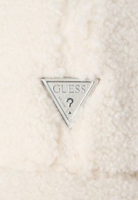 Tissu en fourrure blanche avec une texture douce, présentant un logo en métal triangulaire argenté avec "GUESS" et un point d'interrogation au centre.
