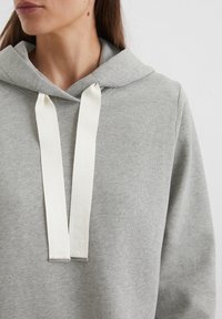Grijze sweatshirt met capuchon, met een gladde textuur en witte koordjes. De koordjes hebben een verhoogde branding.