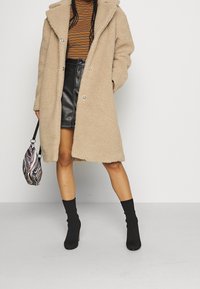 Beiger, flauschiger Mantel mit Kapuze, großen Revers und Knöpfen, kombiniert mit einem schwarzen Lederrock und schwarzen Stiefeletten. Hält eine gestreifte Tasche.