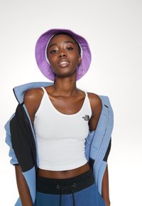 Λευκό κοντό τοπ με μαύρο λογότυπο, συνδυασμένο με ελαφρώς μπλε μπουφάν με φερμουάρ και μωβ καπέλο τύπου bucket. Το μοντέλο φορά σκουλαρίκια κρίκους.